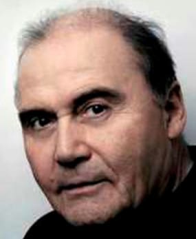 Dramatic, three-quarter-profile, colour, publicity headshot of an unsmiling man with designer stubble and short, receding, dark hair. His dark eyes stare earnestly at the camera from under black eyebrows. He appears to be about to speak.